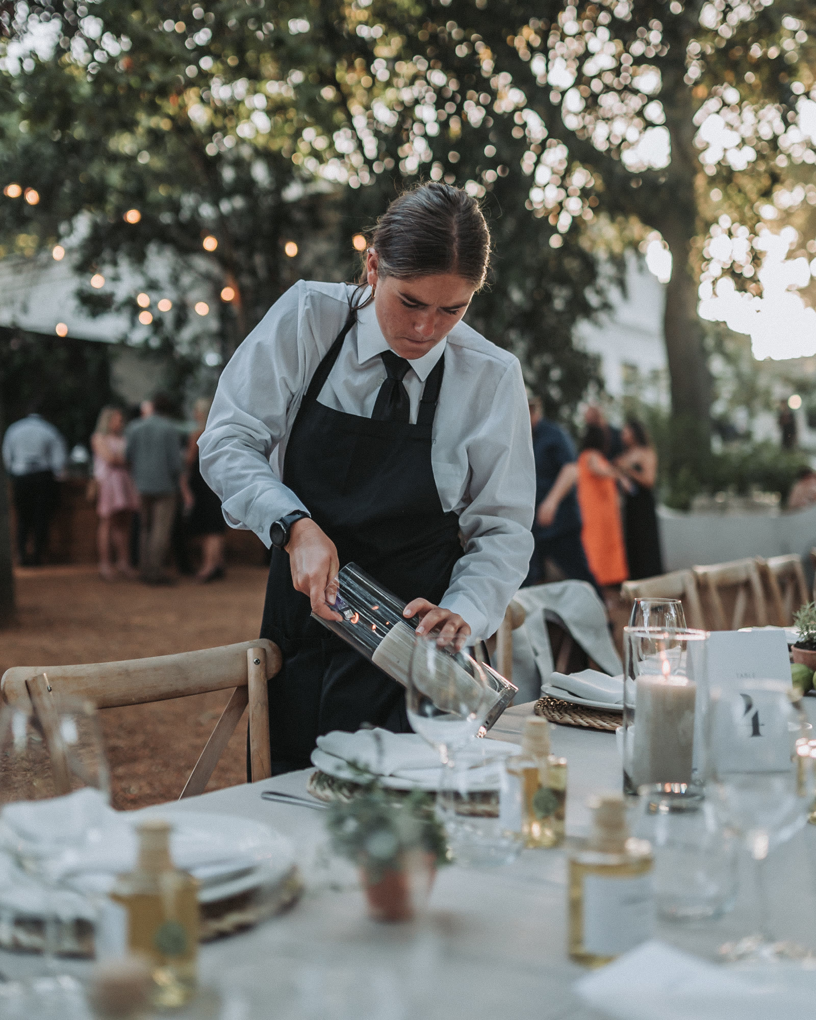Staff lighting candles on an outdoor dinner table under fairy lights